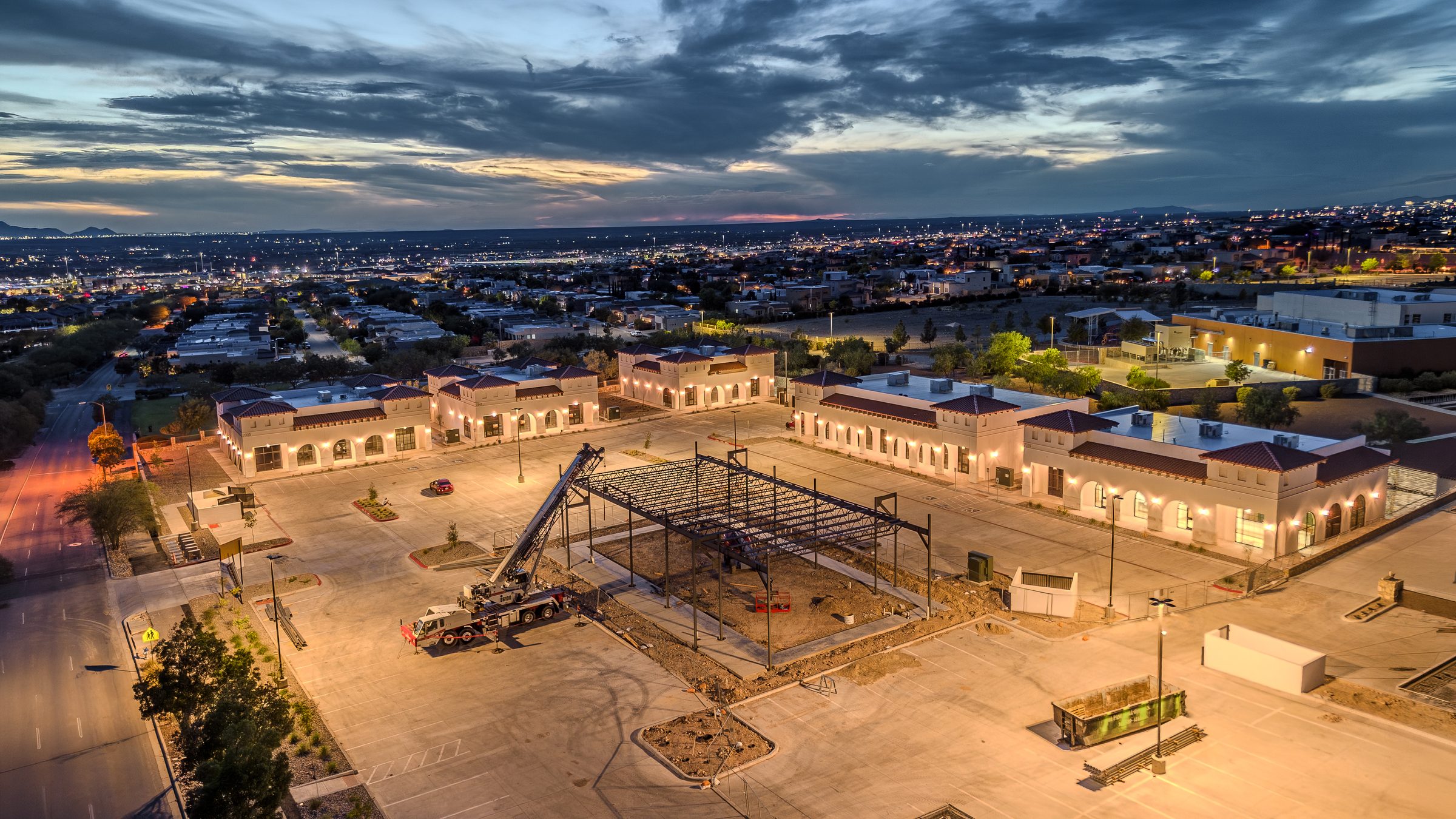 Aerial view at dusk of The Sage at Cimarron medical campus, its Spanish Revival buildings lit against the El Paso valley, with a crane and steel framing for the next building under construction